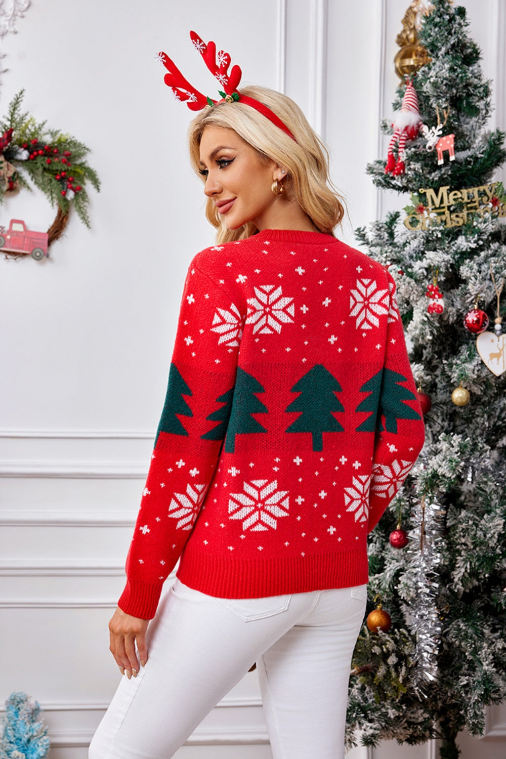 Woman in festive red Christmas sweater with trees and snowflakes, celebrating the holiday season near a decorated Christmas tree.