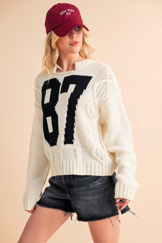 Stylish model in cream cable knit sweater with black number 87, paired with a maroon cap and black denim shorts. Perfect for Christmas!