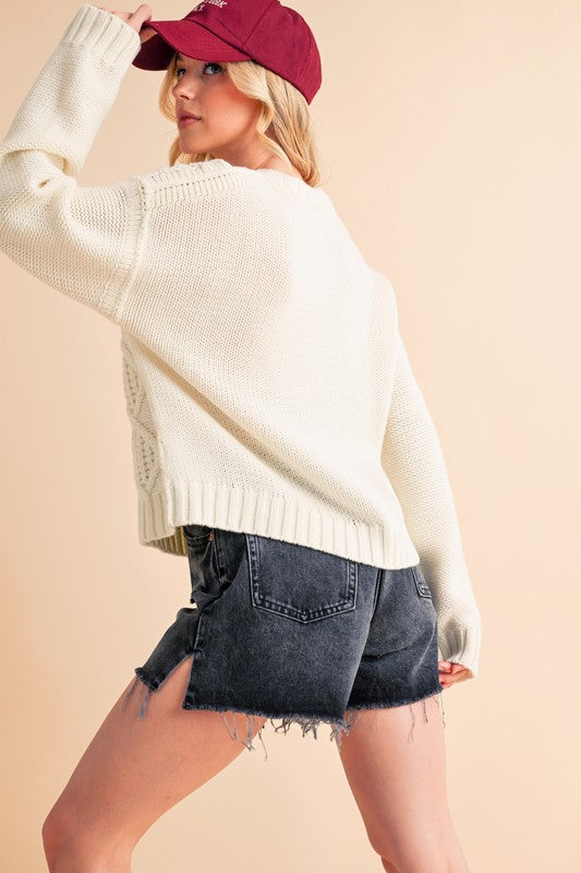 Cozy cream cable knit sweater styled with high-waisted denim shorts and a maroon cap for a trendy look.